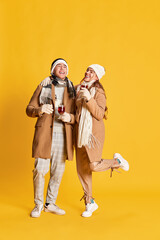 Portrait of stylish young man and woman, smiling coupe in winter clothes walking, drinking mulled wine isolated over yellow background