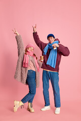 Portrait of cheerful young man and woman in knitted hat and scarf posing, drinking hot cocoa isolated over pink background