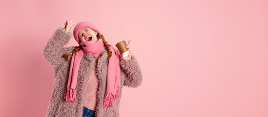 Portrait of emotive young girl in knitted hat, scarf and furry coat, posing with cup isolated over pink background. Hot winter drinks