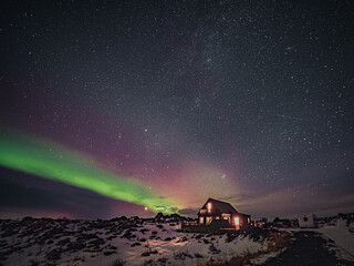 Cozy cottage in winter landscape by night with green and pink aurora borealis