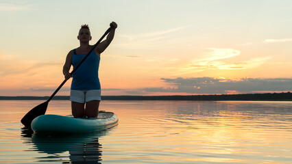 A woman on a kneeling SUP board with an oar at sunset against a golden sky floats in the water of the lake.