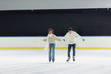 Smiling multiethnic couple in sweaters holding hands while ice skating on rink