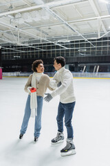 Positive african american woman holding red gift and looking at boyfriend on ice rink