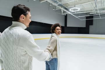 Fototapeta na wymiar Smiling african american woman looking at camera while holding hand of boyfriend on ice rink
