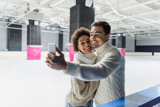 Positive multiethnic couple taking selfie on smartphone on ice rink - Powered by Adobe
