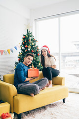 Young beautiful happy Asian woman wearing a Santa Claus hat surprises her boyfriend with gift at home with a Christmas tree in the background. Celebrate the boxing day concept. Photo with copy space.