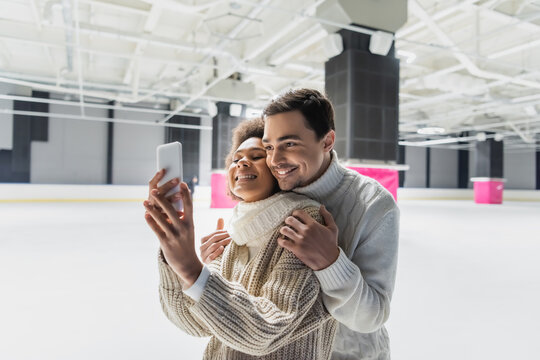 Smiling man hugging african american girlfriend taking selfie on smartphone on ice rink