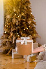 Woman hand holding gift New Year cardboard box with white bow, place for text or logo, with Christmas tree, garlands