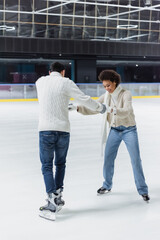 Smiling african american woman holding hands of boyfriend in ice skates on rink