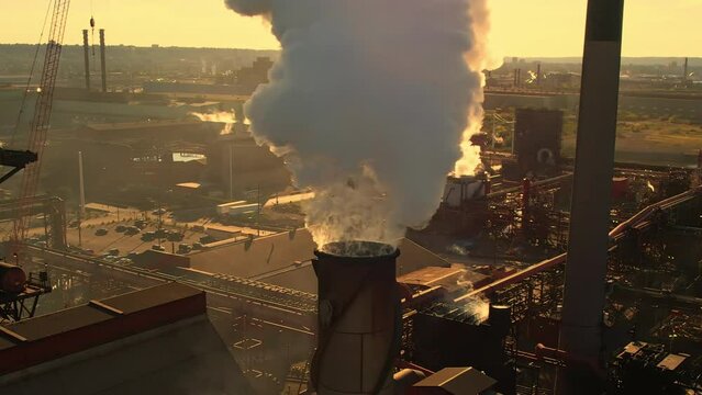 Steel factory pipes during sunset time and golden hour. Sunlight behind thick smoke. Metallurgical plant and smoke cloud is coming from chimney creating greenhouse gases zone layer. Climate change.