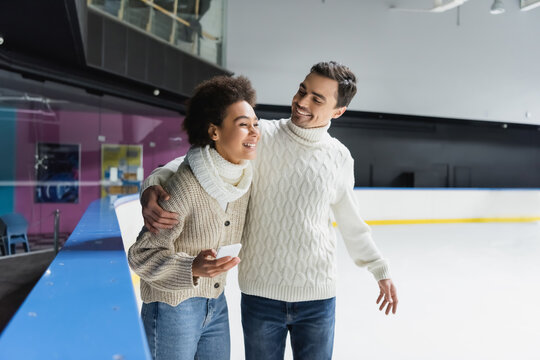Positive man in sweater hugging african american girlfriend with smartphone on ice rink