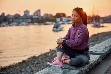 Fototapeta na wymiar Pensive Asian athletic woman relaxing by water at sunset.