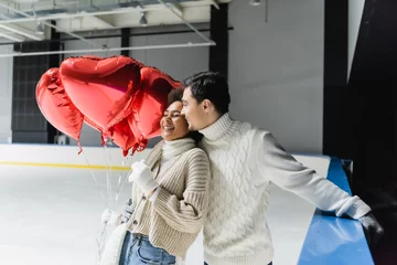 Fotobehang Pleased man in sweater hugging african american girlfriend with heart shaped balloons on ice rink © LIGHTFIELD STUDIOS