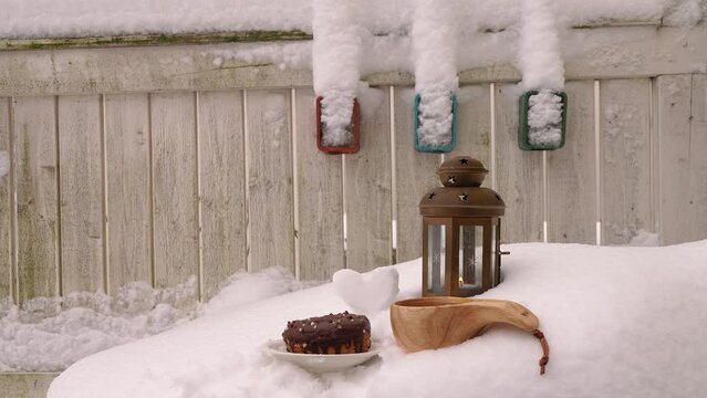 A decorative lantern with a candle, a wooden mug and a chocolate donut in the snow on the background of a wooden fence.