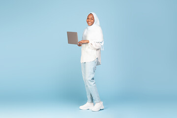 Full length shot of happy black muslim woman in casual wear using laptop computer for online work, blue background
