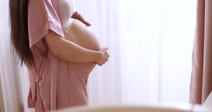 pregnant woman touching belly and standing near window