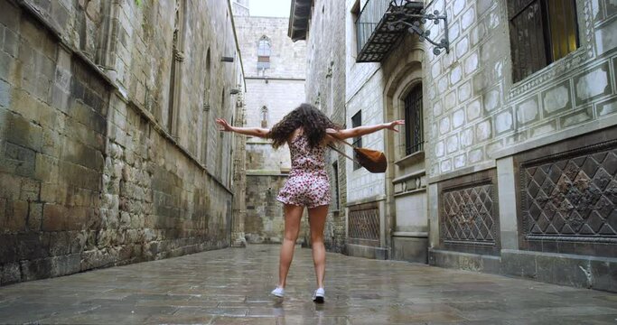 Woman, rain and city for street dancing with happiness, holiday or tourism vacation in Spain. Young dancer girl, happy spin dance and water on road, urban metro and old buildings on travel in Madrid