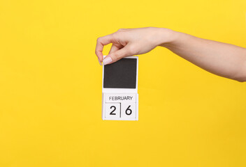 Female hand holding wooden calendar with date February 25 on yellow background