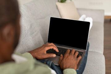 Naklejka premium Busy middle aged african american male on sofa watch video, typing on laptop with blank screen at spare time