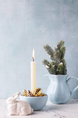 White burning candle, golden decorative cones, white bunnies and branches of Christmas tree in b pitcher on white marble background against blue  textured wall. Rabbit is symbol of 2023..  - 550666787