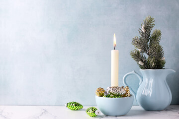 Winter holidays p composition. Green, golden and silver cones decorations, white burning candle and fir tree branches in pitcher  against blue  textured wall. Selective focus. Place for text. - 550666793
