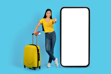 Cheerful young japanese lady in yellow t-shirt with suitcase, tickets show thumb up near huge phone