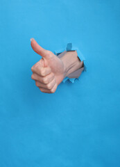 Hand showing thumb up through torn hole of blue paper