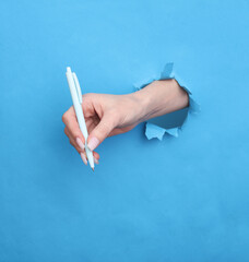 Female hand holds pen through torn hole of blue paper