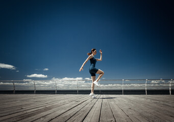 Sporty Caucasian woman practicing jumping up on the embankment at bright sunny day with blue sky and clouds