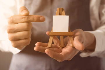 The male artist's hands are holding a miniature easel with a small piece of white paper on it. A creative idea for inviting and discovering art. - obrazy, fototapety, plakaty