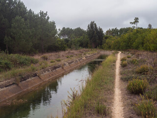 Fototapeta na wymiar Footpath of hiking trail Rota Vicentina along water irrigation canal. Pine trees and green bushes