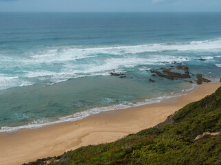 View of empty Praia da Carreagem beach with ocean waves, cliffs and stones, wet golden sand and green vegetation at Rota Vicentina coast, Odemira, Portugal. Cloudy autumn day.