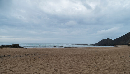 View of empty Praia da Amoreira beach with ocean waves, cliff, stones and golden sand. Rota Vicentina coast, Odemira, Portugal. Cloudy moody autumn day.