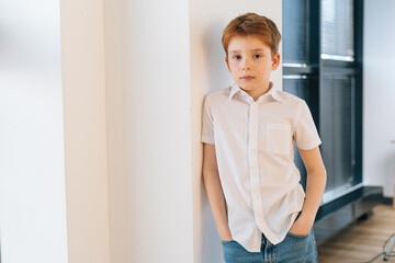 Medium shot portrait of calm handsome child boy model leaning against wall keeping hands in jeans pockets and looking at camera. Confident male kid in stylish outfit posing alone indoors.