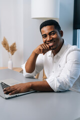 Fototapeta premium Vertical portrait of cheerful African male entrepreneur using laptop and looking at camera while enjoying work sitting at desk in modern office room with light interior, on background of window.