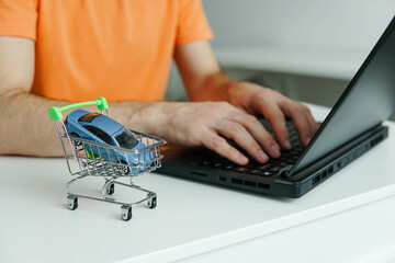 Small shopping cart with miniature car on the table. Man using laptop for byuing new car online.