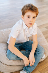 Vertical portrait of handsome child boy looking at camera sitting on pillow on floor in living room with light modern interior. Top view of serious kid posing at home alone, top view.