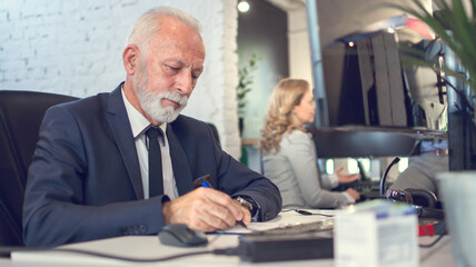 Senior business man wearing formalwear writing on paper in front of computer in office.