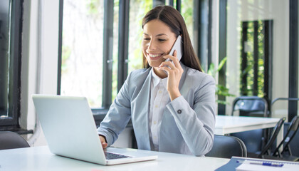 Smiling businesswoman talking on mobile phone and using laptop in office.