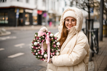 cheerful woman holds wreath of fir branches decorated with glitter balls and plants and bow in hands