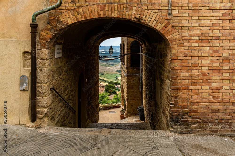 Poster narrow street through an arch between old stone houses of ancient, historical town, volterra, italy