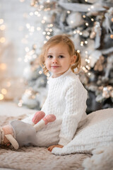 Obraz na płótnie Canvas Very nice charming little girl blonde in white dress sitting on a child's bed and laughs on the background of Christmas trees in bright interior of the house