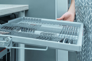 Human hand pull out shelf of dishwasher with clean sparkle cutlery. Dish washing, spoons and forks,...