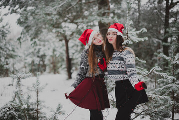 Two young teenage hipster girl friends together.Close up fashion portrait of two sisters hugs and having fun winter time,wearing red santa hats and sweater,best friends couple outdoors, snowy weather