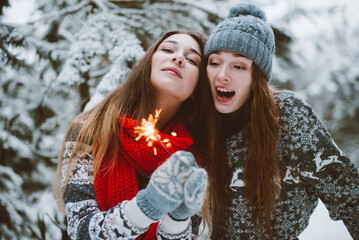 .Close up fashion portrait of two sisters hugs and having fun, holding sparklers in winter time forest, wearing sweaters and scarfs,best friends couple outdoors, snowy weather