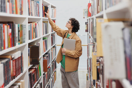 young beautiful woman with curly hair in the library among the shelves with books. a female student conducts scientific work or writes a diploma. a happy confident woman with books in her hands. work