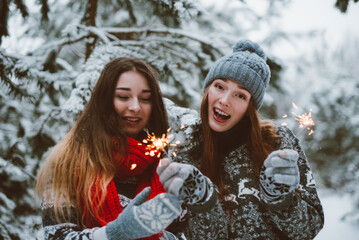 Close up fashion portrait of two sisters hugs and having fun, holding sparklers in winter time forest, wearing sweaters and scarfs,best friends couple outdoors, snowy weather
