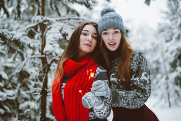 Close up fashion portrait of two sisters hugs and having fun, holding sparklers in winter time forest, wearing sweaters and scarfs,best friends couple outdoors, snowy weather