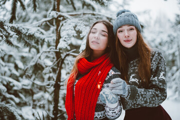 Close up fashion portrait of two sisters hugs and having fun, holding sparklers in winter time forest, wearing sweaters and scarfs,best friends couple outdoors, snowy weather