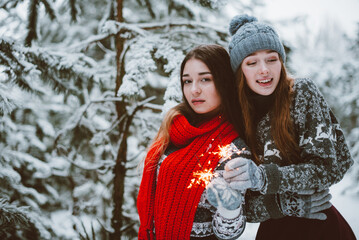 Fototapeta premium Close up fashion portrait of two sisters hugs and having fun, holding sparklers in winter time forest, wearing sweaters and scarfs,best friends couple outdoors, snowy weather